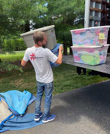 BG Home Mover loading plastic tubs in the back of a moving truck in a driveway with moving blankets on the ground.